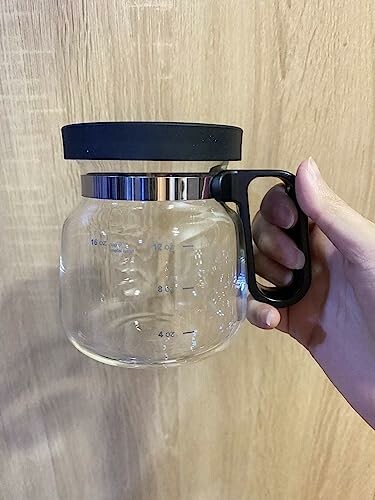 Hand holding a glass coffee carafe with black handle and lid.