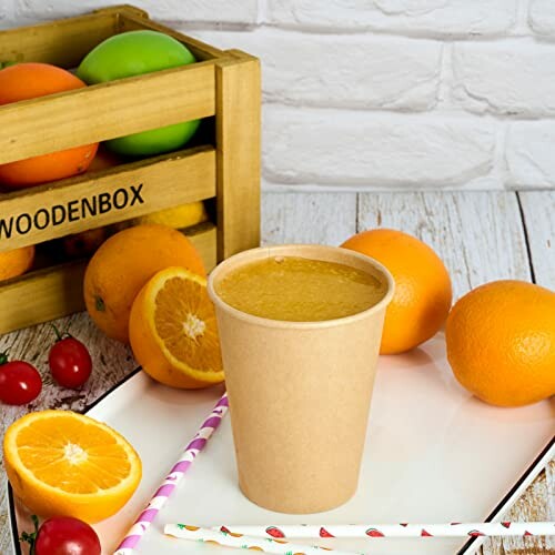 Orange juice in a paper cup surrounded by fresh oranges and fruits.