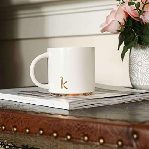 White mug with letter 'k' on magazine beside a vase of pink flowers